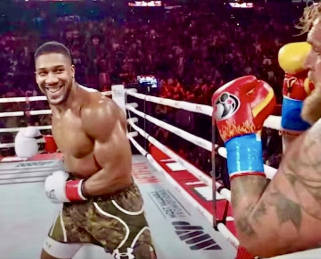 A photograph of two boxers in a professional boxing ring. The boxer on the left is shirtless, muscular, wearing camouflage-patterned shorts with white and red gloves, and is smiling broadly. The boxer on the right has visible tattoos on his arm, is wearing red gloves with blue wrist wraps, and is facing the left boxer. The ring has white ropes, a mat with 'MGM' visible, and a large crowd of spectators in the background, indicating a live event.