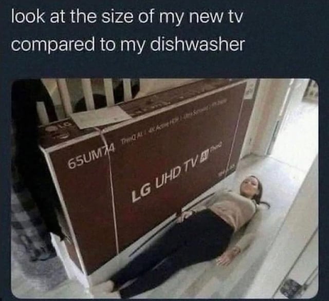 The image shows a large brown LG UHD TV box (model 65UM74) placed on the floor of a room. A person is lying on their back beside the box, stretching out to visually emphasize the TV's size. Above the image, text reads: 'look at the size of my new tv compared to my dishwasher'—a humorous misdirection, as the comparison is actually to the person (not a dishwasher), highlighting the TV's large dimensions. The person appears to be smiling, adding a lighthearted tone to the scene.