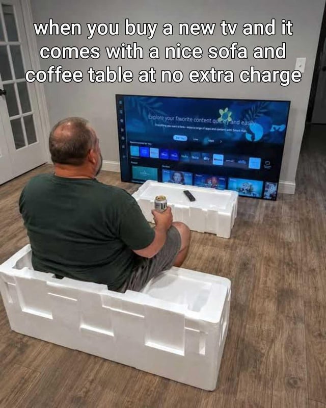 This humorous single-photo meme depicts a middle-aged man viewed from behind, sitting inside a cut-out section of the white styrofoam protective packaging that his new television came in, using a second matching piece of styrofoam packaging as a coffee table to hold his canned beverage and TV remote. The brand new TV is propped directly against the bare wall in front of him, displaying its default setup screen. Overlaid text on the top of the image jokes that the man got a nice sofa and coffee table for free as a bonus with his new TV purchase, leaning into sarcasm about the relatable, lazy/frugal experience of using product packaging as temporary furniture immediately after a large purchase, before acquiring proper home furnishings.