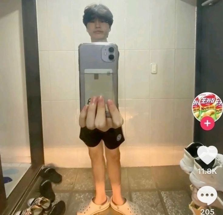 This is a funny bathroom mirror selfie featuring a forced perspective optical illusion. A young man holds his smartphone directly in front of his upper torso, positioned perfectly right below his face. This placement makes the hand holding the phone appear to be attached directly to his neck, creating the false impression that he has an extremely short, tiny body consisting only of legs below the hand holding the phone, with his actual full torso hidden behind the device. The background shows a tiled bathroom, multiple pairs of shoes on the floor, and TikTok interface elements including a like counter, comment counter, and a Korean snack advertisement sticker on the right side of the frame.