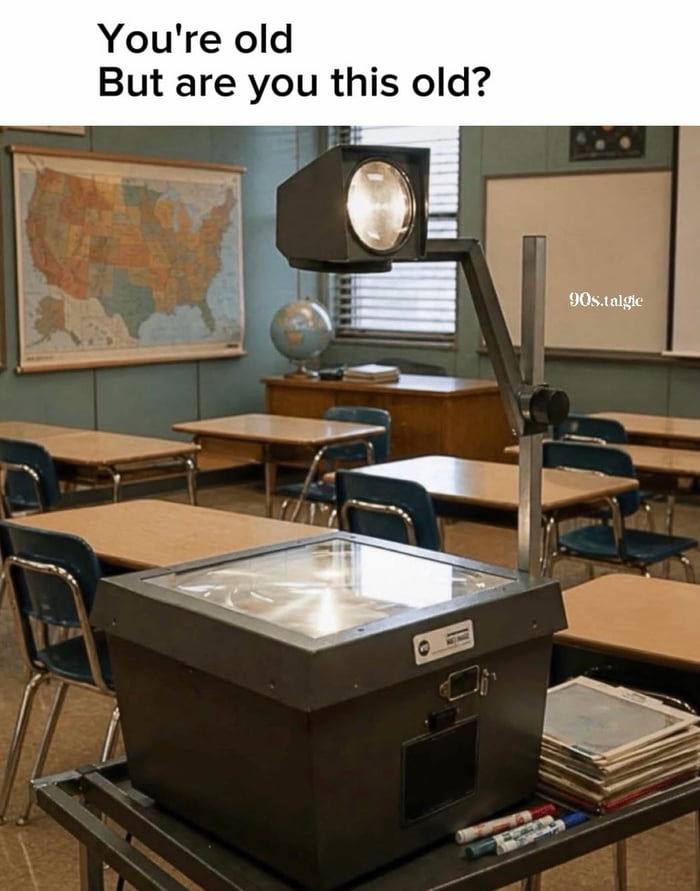 The image displays a vintage overhead projector placed on a rolling cart in a classic late 20th-century North American classroom. The classroom is furnished with wooden and metal student desks, a wall map of the United States, a desktop globe, and a whiteboard in the background. Text at the top of the image reads 'You're old But are you this old?', posing a playful question to viewers to gauge if they are old enough to remember using overhead projectors, devices used to project content from transparent plastic sheets onto walls or screens for class lessons, during their school years. A '90s.talgic' watermark appears on the right side of the image, referencing 1990s nostalgia content.