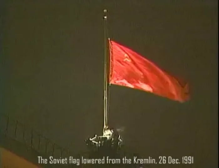 The image depicts the Soviet flag being lowered from the Kremlin in Moscow on December 26, 1991. The red flag with the hammer and sickle emblem is shown mid-descent, fluttering against a dark sky. The base of the flagpole is visible, with part of the Kremlin's architectural structure at the bottom of the frame. A caption at the bottom of the image reads: 'The Soviet flag lowered from the Kremlin, 26 Dec. 1991,' indicating the historical context of the Soviet Union's dissolution.