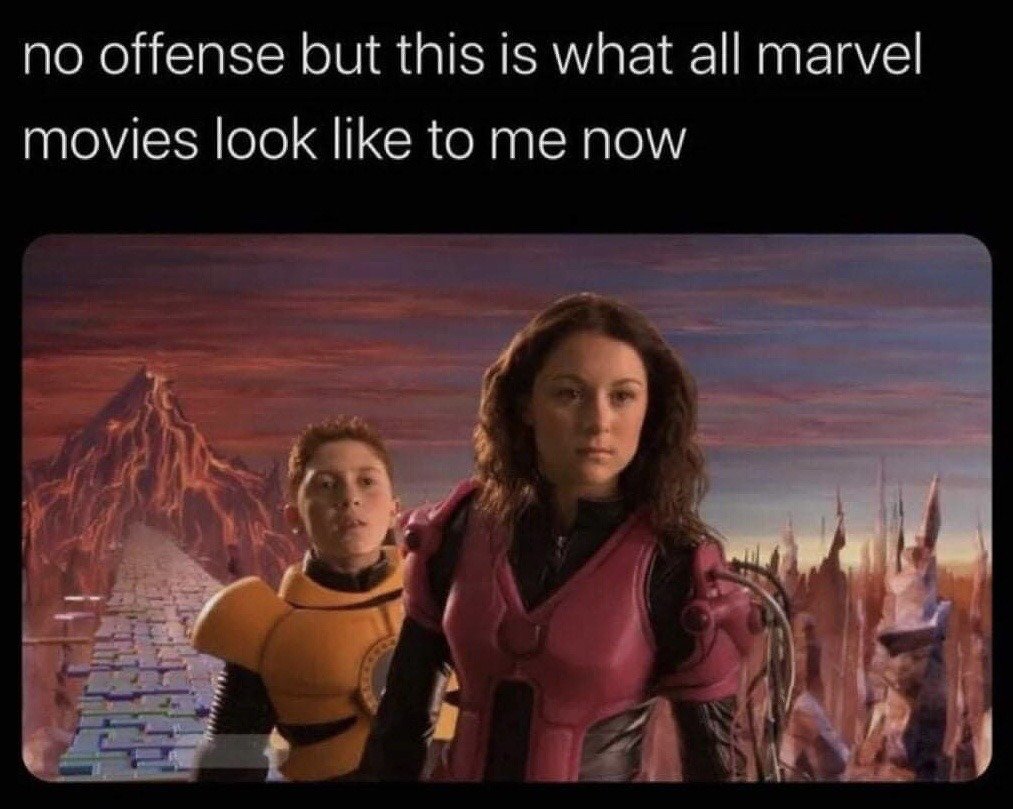 The meme features white text at the top reading "no offense but this is what all marvel movies look like to me now", placed above a still from the 2005 live-action children's fantasy film *The Adventures of Sharkboy and Lavagirl*. The still shows the character Lavagirl in a pink futuristic suit standing in the foreground with a neutral serious expression, and the protagonist Max, wearing a yellow suit, standing behind her with a wide-eyed surprised look. They are positioned in front of a highly saturated, stylized CGI fantasy landscape with colorful skies, jagged rock formations, and blocky paved ground. The joke satirizes common criticisms of recent Marvel Cinematic Universe films, which are often panned for overusing overly bright, cheap-looking CGI and generic sci-fi/fantasy visual aesthetics by comparing them to the intentionally whimsical, lower-budget CGI of the 2005 children's movie.