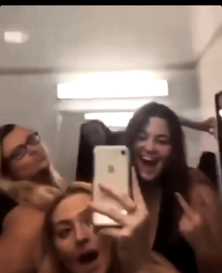 This is a low-quality candid mirror selfie captured inside a residential bathroom. Three young women are gathered in the foreground posing for the photo: the woman on the left wears glasses and has long blonde hair, holding a playful pose; the woman in the lower center has her mouth open in an expression of excited surprise; the woman on the right is laughing broadly while holding an older model white iPhone to take the mirror selfie. The central comedic detail is in the background: over the top of a dark shower curtain, the head and raised arm of a fourth person are partially visible, appearing to hide in the shower area completely unnoticed by the three women taking the photo. The meme is paired with the title 'Too much to bear', referencing the absurd, shocking nature of discovering a hidden person in your bathroom while taking a casual selfie.