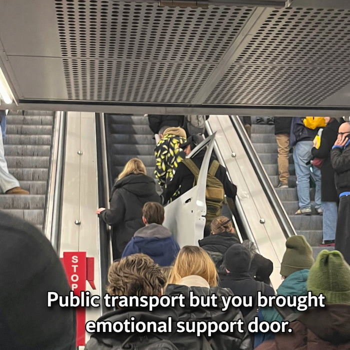 A photograph of a busy escalator in a public transport station, likely a subway or train station. The escalator is crowded with commuters wearing winter clothing (coats, hats). In the center of the frame, a person is prominently carrying a large, white door on their back, as if transporting it. At the bottom of the image, a caption reads: 'Public transport but you brought emotional support door,' which humorously recontextualizes the door as an 'emotional support' item, a playful twist on the concept of emotional support animals or objects. The scene blends everyday urban life with an absurd, whimsical element, creating a funny and relatable meme about public transit experiences.