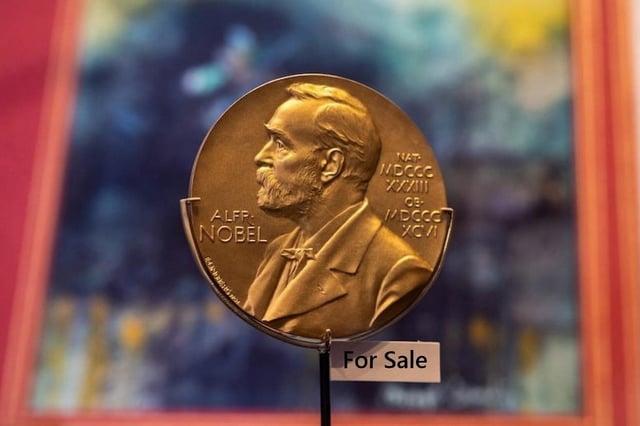 A photograph of a gold-colored Nobel Prize medal featuring the portrait of Alfred Nobel, with inscriptions including 'ALFR. NOBEL' visible. The medal is mounted, and a small sign reading 'For Sale' is placed in front of it. The background is blurred with colorful, indistinct patterns. The image humorously suggests the prestigious Nobel Peace Prize is being offered for sale, creating a satirical commentary on the award's perceived value or integrity.