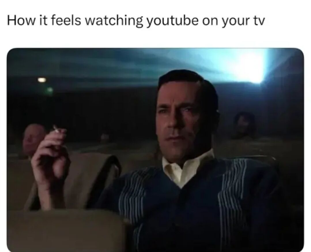 This is a relatable humor meme that describes the common experience of watching YouTube on a home television. The meme uses a still frame from the TV series *Mad Men* showing the character Don Draper sitting alone in a dark, nearly empty movie theater, holding a cigarette with a calm, contemplative expression. The joke plays on how watching YouTube alone on your big TV at home often feels like being alone in a large dark theater, a quiet solo experience different from traditional group TV watching.