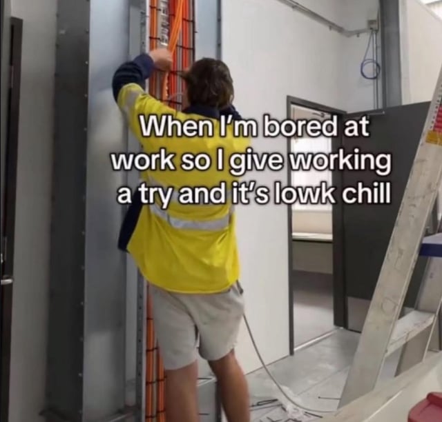 This meme features a real-life worker in a yellow high-visibility shirt standing and working on an electrical utility panel filled with orange wiring. Overlaid on the image is the text: 'When I'm bored at work so I give working a try and it's lowk chill'. The joke plays on the relatable experience of feeling bored at work, then discovering that engaging in the actual work can be surprisingly low-key enjoyable or relaxing. The term 'lowk' is internet slang shorthand for 'low key'.
