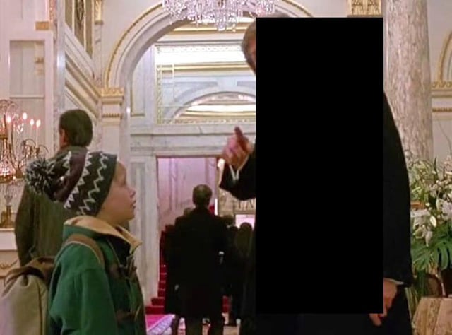 A screenshot from the 1990 film 'Home Alone' featuring the main character Kevin McCallister (played by Macaulay Culkin) in a grand, ornately decorated lobby with marble columns, chandeliers, and red carpet. Kevin, wearing a green jacket and a patterned beanie, looks up at an adult male character whose face has been edited out with a solid black rectangle. The man is dressed in a dark suit and appears to be pointing his finger towards Kevin. Several other people are visible in the background, walking or standing, contributing to the busy, formal atmosphere of the scene.