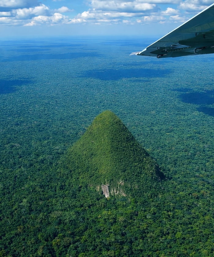 An aerial photograph capturing a vast expanse of dense, lush green forest. Dominating the center of the image is a striking, pyramid-shaped hill covered in thick vegetation, with a small exposed rock face visible near its base. The forest stretches endlessly in all directions, creating a sea of green that blends into the horizon under a clear sky with scattered white clouds. A portion of an airplane wing is visible in the top right corner, confirming the aerial vantage point from which the photo was taken. The scene conveys a sense of natural grandeur and untouched wilderness.