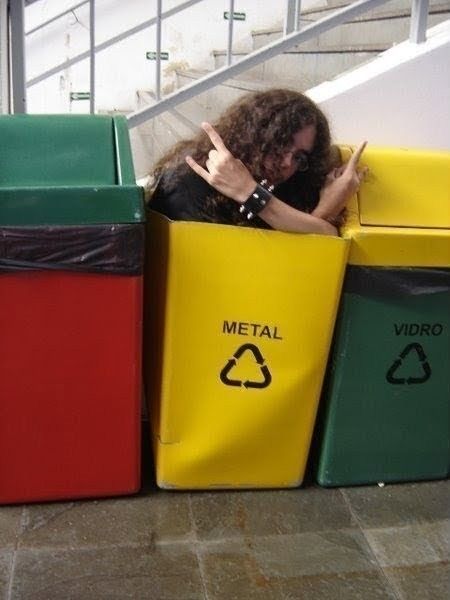 The image features a person with long curly hair and a studded wristband, stereotypical of heavy metal subculture, sitting inside a bright yellow recycling bin that is explicitly labeled 'METAL' for metal waste recycling. The person leans out of the bin making the iconic 'devil horns' rock hand gesture with both hands. Other recycling bins are placed beside the 'METAL' bin, including a green bin labeled 'VIDRO' (Portuguese for glass), and a public area with concrete stairs is visible in the background. The joke relies on a pun playing on the double meaning of the word 'metal': the material type the bin is intended for, and the heavy metal music subculture the person belongs to.