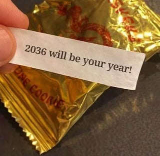 A close-up image of a hand holding a white fortune cookie slip with the text '2036 will be your year!' in black font. In the background, a gold-colored fortune cookie wrapper is partially visible, with the text 'FORTUNE COOKIE' printed in red. The image humorously or optimistically references a distant future year as a 'good year' prediction, playing on the typical fortune cookie trope of vague, positive forecasts.
