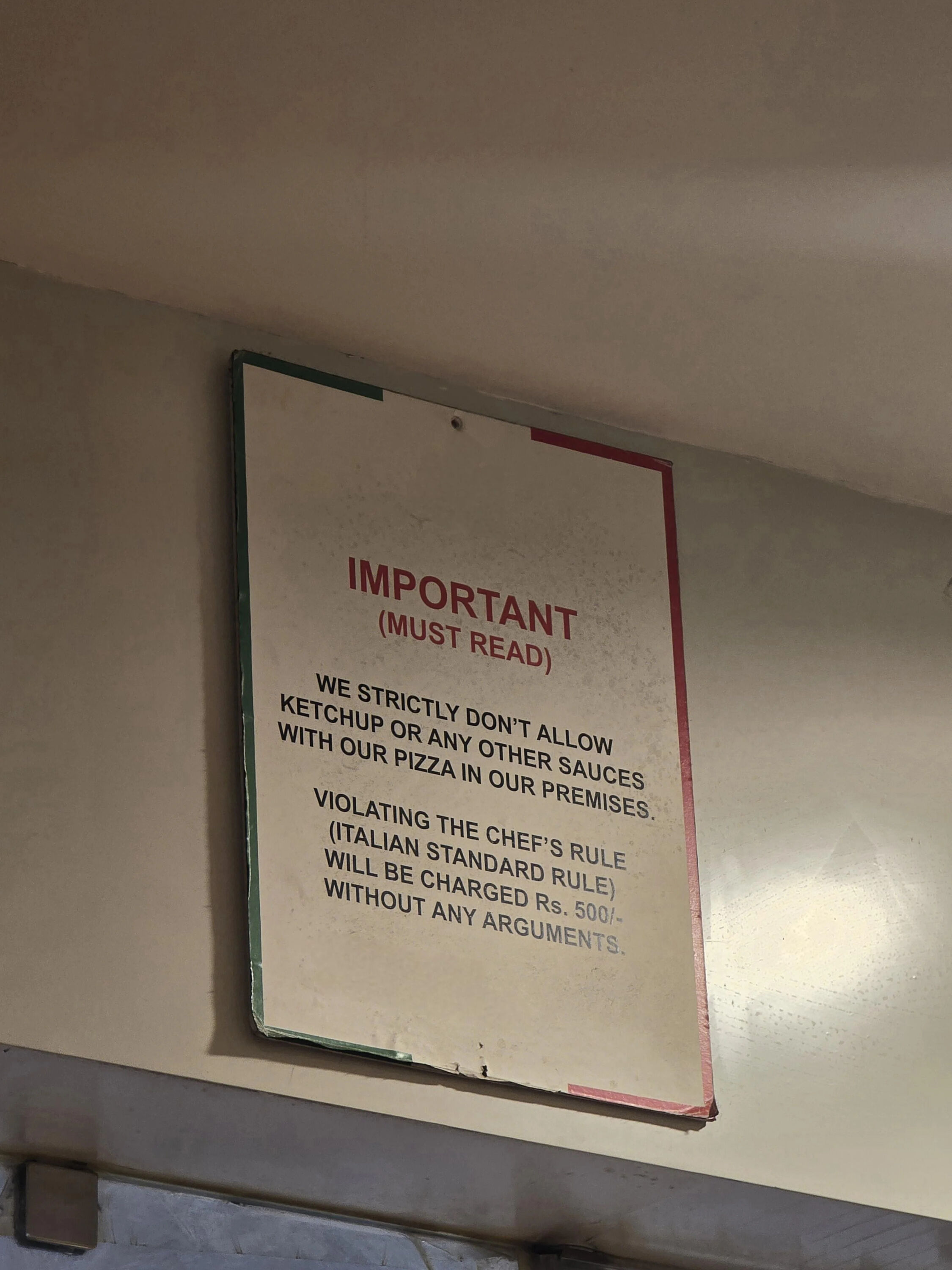 A photograph of a rectangular sign mounted on a beige wall, likely in a pizza restaurant. The sign has a slightly worn appearance with a green and red border. At the top, bold red text reads 'IMPORTANT (MUST READ)'. Below, black text states the establishment's strict policy: 'WE STRICTLY DON'T ALLOW KETCHUP OR ANY OTHER SAUCES WITH OUR PIZZA IN OUR PREMISES. VIOLATING THE CHEF'S RULE (ITALIAN STANDARD RULE) WILL BE CHARGED Rs. 500/- WITHOUT ANY ARGUMENTS.' The sign shows minor wear on the edges and surface, indicating it has been displayed for some time.