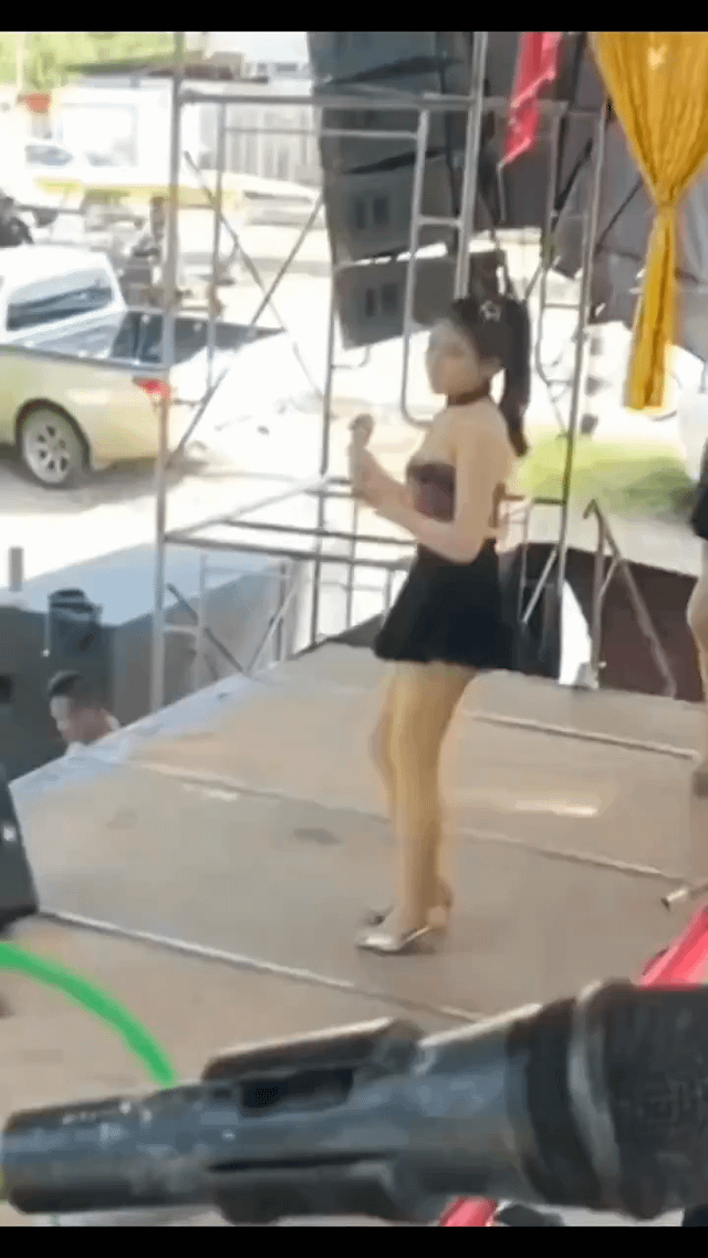 The image captures an outdoor live performance scene on a simple metal-framed stage. A female performer, dressed in a black halter short dress, gold sheer tights, and shiny flat shoes with her dark hair tied back, stands on the stage holding a microphone, seemingly in the middle of a performance. In the lower foreground, a large microphone partially obscures the view. To the left, just off-stage, a man is seated, and the camera's perspective creates an accidental optical illusion: the alignment of the performer's lower body with the man's upper body makes it appear as if the performer has an abnormally short torso or that her legs are not her own, creating a humorous, misleading visual. The background includes a yellow pickup truck, stage scaffolding with large speakers, and an open outdoor area with other vehicles and structures visible in the distance.