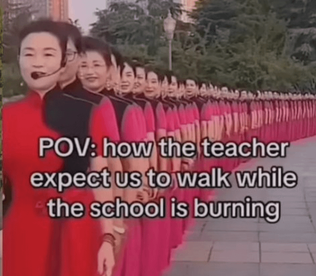 The image features a long line of individuals, likely women, dressed in coordinated red and black traditional-style dresses walking in perfect synchronization on a paved path with greenery and buildings in the background. Overlaid text reads 'POV: how the teacher expect us to walk while the school is burning,' creating a humorous contrast between the strict, orderly behavior expected by teachers and the chaotic scenario of a burning school, mocking overly rigid rules in stressful situations.