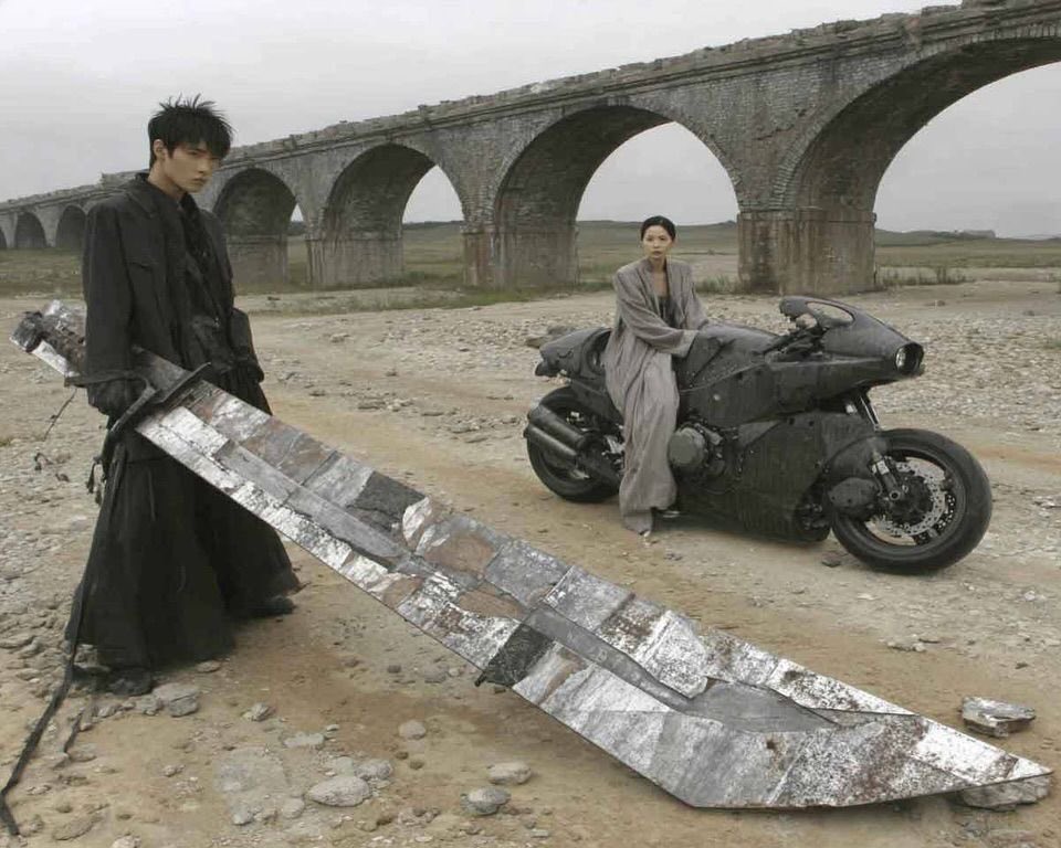 The image is set in a desolate, rocky wasteland under an overcast sky, with a long, weathered multi-arch stone aqueduct bridge spanning the background. On the left, a young man with spiky dark hair wearing a long loose black coat stands holding the hilt of an extremely large, oversized broad sword made of patched scrap metal that rests on the ground and extends nearly the full length of the frame. On the right, a woman with short dark hair wearing a loose flowing light grey robe sits atop a matte black, futuristic modified heavy motorcycle, looking toward the camera. The overall aesthetic is gritty, dystopian, and incorporates dark fantasy and post-apocalyptic design elements.