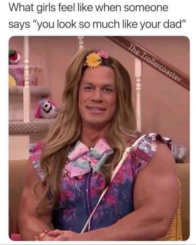 A humorous meme depicting a person with a muscular build wearing feminine attire (a floral dress, large pastel bow, and a yellow flower in long blonde hair) sitting in a domestic setting. The text overlay reads, 'What girls feel like when someone says "you look so much like your dad"', using the contrast between the typically masculine physique and feminine clothing to exaggerate the awkward or amused reaction to being compared to one's father.