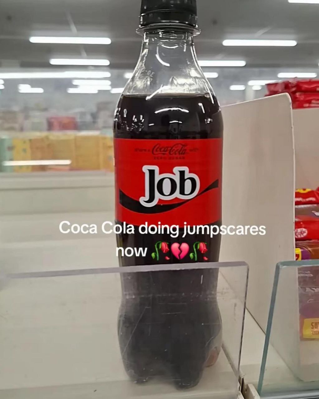 A photograph of a Coca-Cola bottle placed on a store shelf in a supermarket. The bottle's iconic red label has been digitally altered, replacing the 'Coca-Cola' brand name with the word 'Job' while retaining the original font style and logo placement. Overlaid text on the image reads 'Coca Cola doing jumpscares now' accompanied by a broken heart emoji (💔) and a rose emoji (🌹). The background shows blurred supermarket shelves with various products, emphasizing the bottle as the focal point.