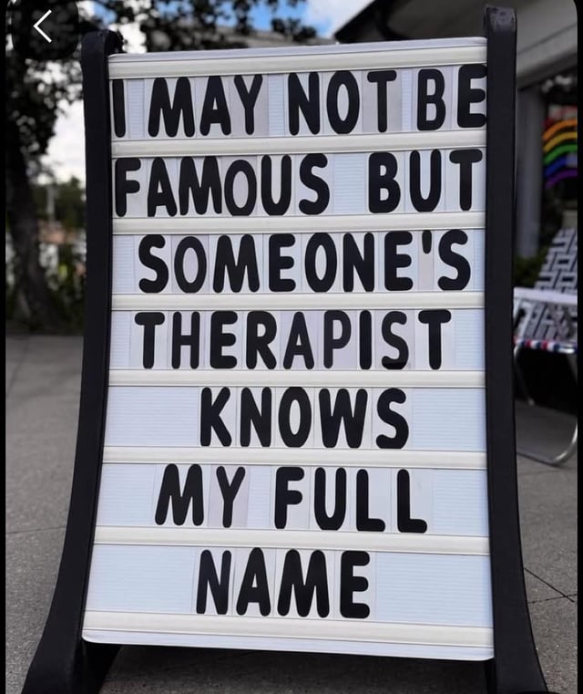 This image is a photograph of a freestanding A-frame letter board sign placed on an outdoor concrete sidewalk. The sign displays large black sans-serif text spread across seven lines that reads: 'I MAY NOT BE FAMOUS BUT SOMEONE'S THERAPIST KNOWS MY FULL NAME'. The background is softly blurred, showing green trees, a building exterior, a small rainbow pride decoration, and a patterned folding chair on the right side. The joke is a lighthearted, self-deprecating quip that plays on the concept of fame: instead of being widely known for positive accomplishments, the speaker is known well enough by someone that their full name is mentioned in another person's therapy sessions, implying they have caused enough stress, drama, or emotional impact to be discussed in a mental health context.