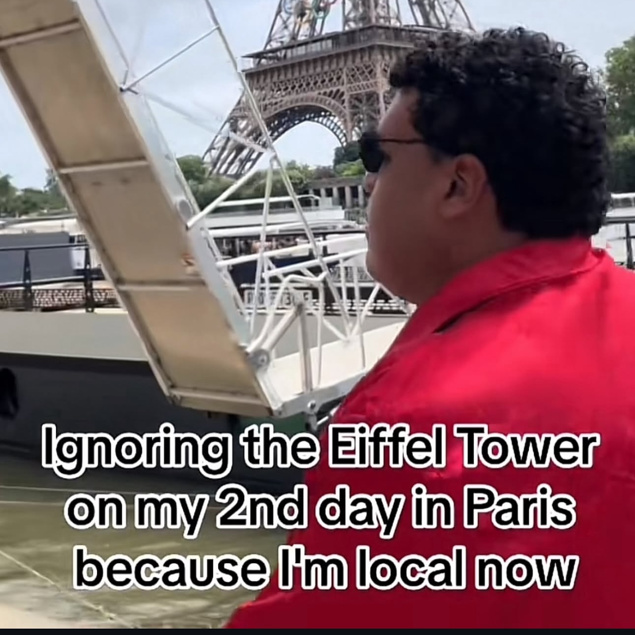 This meme image features a man with curly dark hair, wearing black sunglasses and a bright red jacket, riding a boat on the Seine River in Paris. The iconic Eiffel Tower is clearly visible in the background behind him, but he is looking away from the landmark, appearing completely uninterested in it. White text with a black outline is overlaid at the bottom of the image, reading 'Ignoring the Eiffel Tower on my 2nd day in Paris because I'm local now'. The joke pokes fun at the common relatable behavior of tourists who adopt a casual, 'local' attitude towards famous landmarks after an extremely short period of time visiting a popular destination.