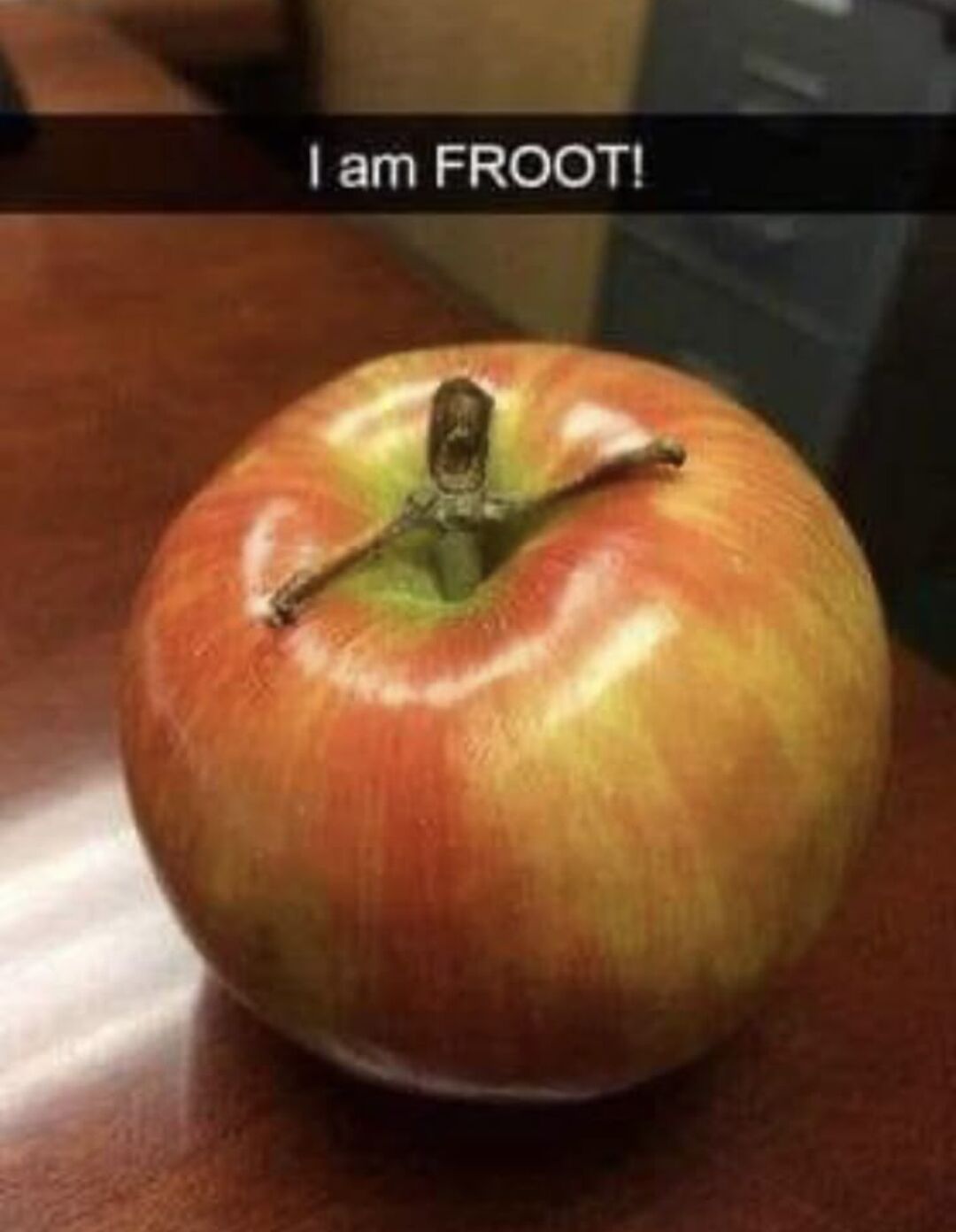 The meme features a photograph of a red and yellow apple resting on a polished wooden table surface. The apple has its main stem plus a smaller extra twig attached to the stem base, arranged in such a way that they look like a tiny person standing on top of the apple with one arm outstretched, creating accidental anthropomorphism. A black bar at the top of the image has white text that reads 'I am FROOT!', a playful misspelling of the word 'fruit'. The humor comes from the personified appearance of the apple paired with its silly, grammatically incorrect declaration of its identity as a fruit.