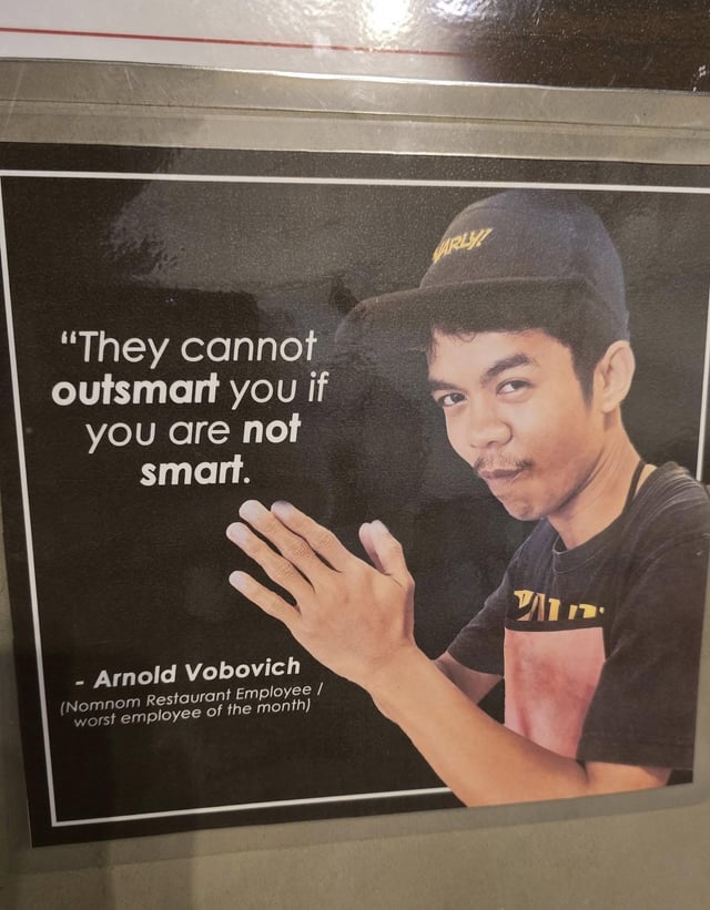 A framed poster featuring a photograph of Arnold Vobovich, identified as a 'Normnom Restaurant Employee' and 'worst employee of the month.' Vobovich is wearing a black cap with yellow text and a black shirt with a red and yellow design. He is making a hand gesture with palms facing upward, as if explaining or dismissing something. The poster has a black background with white text displaying the quote: 'They cannot outsmart you if you are not smart.' The attribution below the quote lists his name, occupation, and 'worst employee' title. The overall design mimics an official employee recognition poster but with a sarcastic twist.
