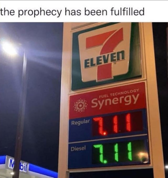 This is a nighttime photo of a gas station price sign. At the top of the sign, the 7-Eleven convenience store logo is displayed. Below the logo, branding for 'Synergy Fuel Technology' is visible, followed by digital price readouts: regular gasoline is priced at 7.11 in red text, and diesel fuel is also priced at 7.11 in green text. Above the photo, a text caption reads 'the prophecy has been fulfilled'. The humor stems from the perfect coincidence that both fuel prices match the numerical namesake of the 7-Eleven brand, framed as a long-awaited prediction coming true. A partial Mobil gas station sign is visible in the bottom left of the frame.