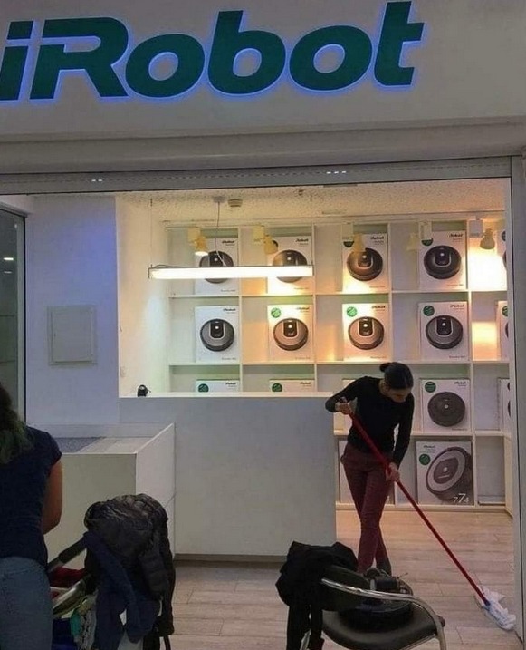 This image captures a highly ironic scene at an iRobot retail store, which specializes in selling Roomba automated robotic vacuum cleaners designed to clean floors without manual labor. Dozens of boxed Roomba units are clearly displayed on built-in shelves across the back wall of the store. The central humorous element is a female store employee who is manually mopping the wood-look floor of the store with a standard mop, rather than using the automated cleaning products that the brand sells to customers. A second woman standing next to a stroller is partially visible at the left edge of the frame, and a chair with coats and personal items is visible in the right foreground.