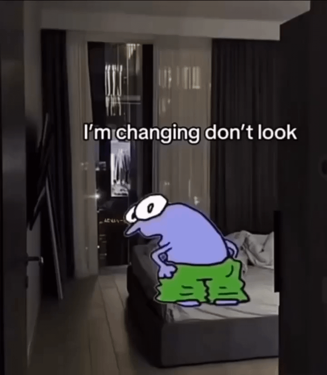 The image is shot from a first-person perspective, looking through an open doorway into a dimly lit modern bedroom with a large bed, dark curtains, and a view of city lights through the window. A crudely drawn purple cartoon character with large wide eyes, a long pointed nose, and extremely baggy oversized green pants is placed on the bed, appearing to be pulling his pants up. Overlaid white text with a black outline at the top of the image reads 'I'm changing don't look'. The humor comes from the absurdity of the character's request: his pants are already pulled all the way up to his chest, covering his entire body completely, so there is nothing to see, framing the viewer who is peeking through the door as a creep, which aligns with the original post title 'Yall are creep'.