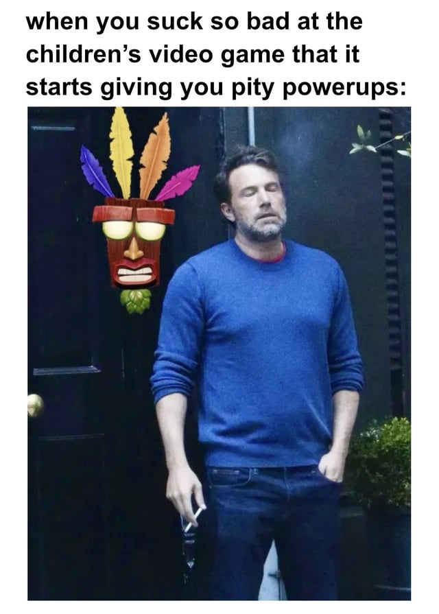 The meme consists of a top text overlay reading: 'when you suck so bad at the children's video game that it starts giving you pity powerups:' followed by a photograph of Ben Affleck. Affleck is standing outdoors against a dark wall, wearing a blue sweater and holding sunglasses in his right hand, with a tired, dejected expression. To his left is a colorful Tiki mask with purple, yellow, and pink feathers, a red and brown wooden face, and a green leaf at the bottom. The combination humorously uses Affleck's sad demeanor to personify the embarrassment or frustration of needing pity mechanics in a simple children's video game, creating a relatable and funny reaction image.