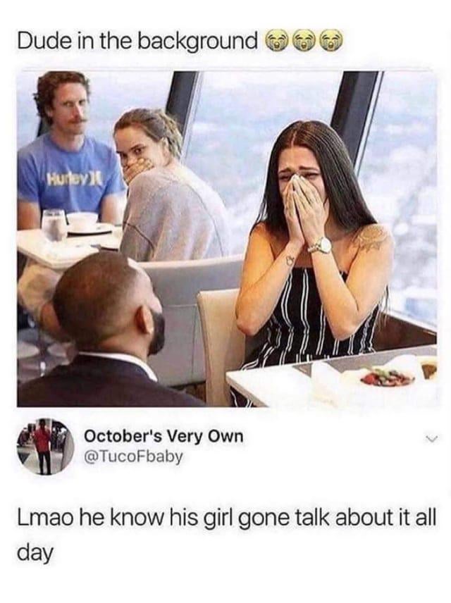 The image is a screenshot of a viral social media meme. Text at the top reads 'Dude in the background' followed by three laughing-crying emojis. The underlying photograph is taken inside a high-rise restaurant with large windows showing a city skyline. In the foreground, a woman with long dark hair wearing a black and white striped dress sits at a table across from a man in a suit (seen from behind), crying into her hands, implying she is having a difficult conversation or argument during their date. In the background booth, a blonde woman covers her mouth laughing at the scene, while her male companion, wearing a blue Hurley t-shirt, looks directly at the crying couple with a smug, amused expression. Below the photo, the original poster's text reads 'Lmao he know his girl gone talk about it all day', joking that the man in the background is aware his girlfriend will be discussing the public drama of the crying couple nonstop after their meal.