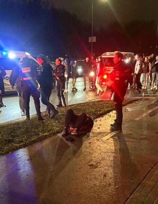 A nighttime scene showing an emergency situation with multiple emergency personnel (likely police or paramedics) attending to a person sitting on the ground near a road. Several vehicles with flashing lights (blue and red) are present, illuminating the area. A crowd of onlookers stands in the background, observing the scene. The ground appears wet, possibly from rain, and the atmosphere is tense.