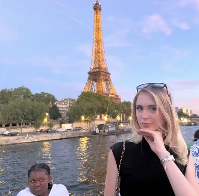 The photo is captured from a river cruise on the Seine in Paris at dusk, with the lit-up Eiffel Tower clearly visible in the background against a pastel evening sky. A blonde woman in a black top poses intentionally in the foreground, resting her hand under her chin and looking at the camera for a standard tourist photo. The humor comes from a young Black girl partially visible in the lower left corner, who is unintentionally in frame, making a deadpan, unimpressed side-eye expression that steals focus and creates a hilarious, unexpected contrast to the posed tourist shot.