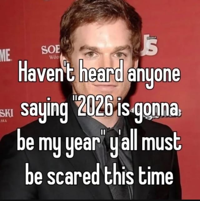 The image features a man in a black suit with a dark shirt, standing against a red background with partially visible logos (including 'US' and 'SOE'). Overlaid in large white text is the caption: 'Haven't heard anyone saying "2026 is gonna be my year" y'all must be scared this time'. The man has a neutral, serious expression and is facing directly toward the camera. The meme humorously observes the absence of people claiming 2026 will be their year, implying past disappointment with similar annual declarations.