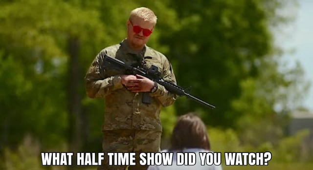An outdoor image showing a person in military camouflage uniform standing and holding a rifle, with green trees in the background. A partial view of another individual with brown hair is visible in the foreground. At the bottom of the image, bold uppercase white text with a black outline reads: 'WHAT HALF TIME SHOW DID YOU WATCH?'