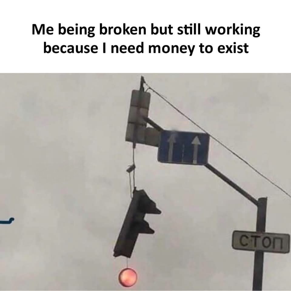 This is a single panel relatable meme. Text at the top of the image reads 'Me being broken but still working because I need money to exist'. Below the text is a photograph of a heavily damaged traffic light hanging loosely from its mounting post by thin wires, visibly broken and nearly detached, but its red indicator light is still illuminated and functional. A blue directional road sign with two straight ahead arrows and a Cyrillic 'СТОП' (stop) sign are also mounted on the same pole, against a grey overcast sky. The humor comes from the comparison between the broken but still operating traffic light and burnt out people who continue working despite struggling, because they need income to cover basic living costs.