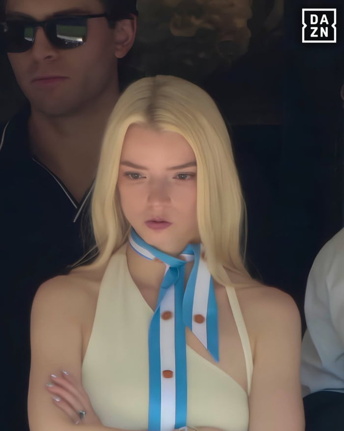 This meme features a candid photograph of actress Anya Taylor-Joy sitting with her arms crossed, wearing a pale yellow halter top and a blue-and-white striped neck scarf, with a clearly disappointed, unamused, gloomy facial expression. A man in sunglasses is partially visible behind her, and a DAZN (sports media platform) watermark is present in the top right corner. The title 'No Joy tonight' is a pun playing on Anya Taylor-Joy's last name 'Joy', referencing her unhappy expression to communicate that the evening or event is not fun, disappointing, or has failed to meet expectations.
