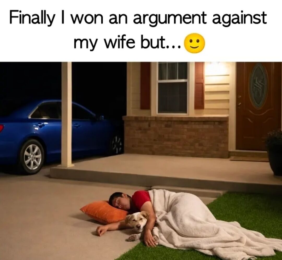 The meme consists of two parts: top text and a photograph. The top text reads 'Finally I won an argument against my wife but... 😊' with a smiling emoji. Below the text is a nighttime photograph showing a man lying on a driveway next to a house. He is covered with a white blanket, resting his head on an orange pillow, and gently holding a dog that is lying beside him. A blue car is parked in the driveway to the left, and the house has a lit porch light, brick accents, and a wooden front door. The scene humorously implies the man 'won the argument' but was locked out, yet the peaceful setting with the dog adds a wholesome twist.