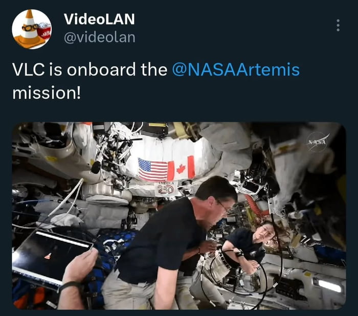 This image is a screenshot of a tweet from VideoLAN's official account, showing the iconic orange cone logo of VLC Media Player displayed on a laptop screen inside the NASA Artemis mission spacecraft in space. Two astronauts can be seen working inside the cramped space capsule, with the flags of the United States and Canada visible on the wall behind them, and a NASA watermark in the corner of the photograph. The joke here is that the famous VLC cone logo is literally located 'in space', matching the meme title.