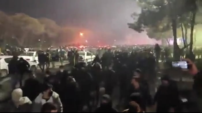 A nighttime photograph depicting a large crowd of people gathered outdoors. The scene is dimly lit, with trees visible on the right side and several parked cars on the left. In the distance, a prominent red light is visible, potentially from a fire or emergency vehicle. Many individuals in the crowd appear to be standing or moving, with some holding up mobile phones, likely recording the event. The atmosphere suggests a significant public gathering, possibly a demonstration or protest, given the size of the crowd and the nighttime context.