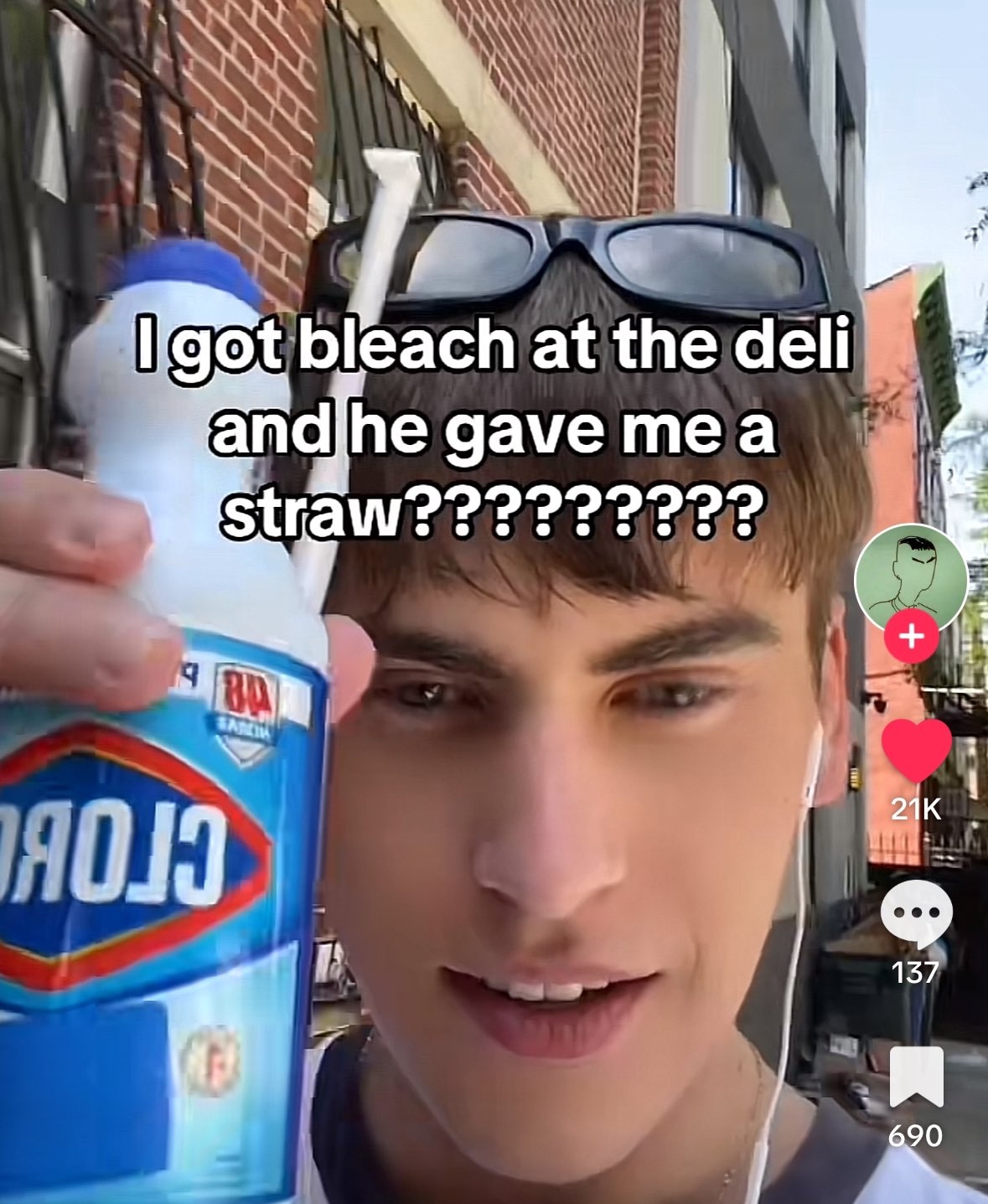 This is a TikTok screenshot of a young man standing on a residential street lined with brick buildings. He holds a bottle of Clorox bleach up to the camera, with a disposable plastic straw inserted into the top of the bottle. He has sunglasses resting on his head, earbuds in, and a confused, amused expression. Overlaid white text on the image reads 'I got bleach at the deli and he gave me a straw??????????'. TikTok interface elements are visible on the right side, showing 21K likes, 137 comments, and 690 saves. The humor comes from the absurd, dangerous mistake of the deli worker giving a straw for a toxic cleaning product, implying they mistook the bleach bottle for an edible beverage while working on autopilot.