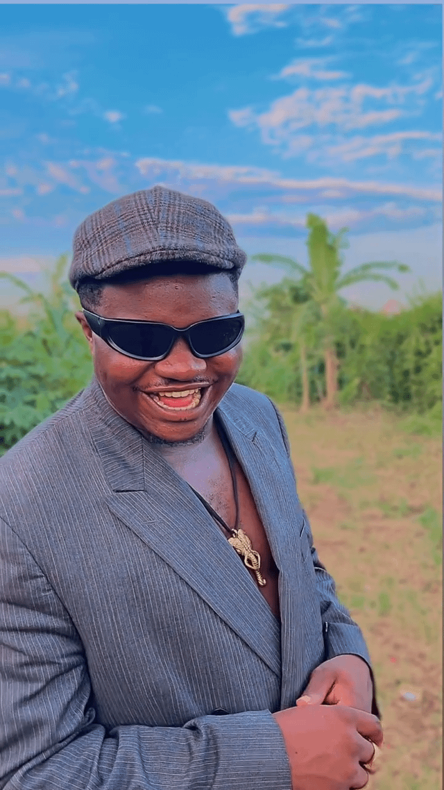 A man stands outdoors in a rural setting wearing a gray pinstriped suit jacket, matching gray cap, black sunglasses, and a gold elephant pendant necklace. He is smiling broadly at the camera. The background features green vegetation, palm trees, and a blue sky with scattered clouds, suggesting a warm, sunny day in a countryside location.
