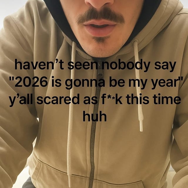 A close-up image of a person wearing a beige hoodie with a black inner lining. The individual is positioned such that their upper torso and part of their face (focused on the mouth and mustache) are visible. Overlaid on the image is bold black text that reads: 'haven’t seen nobody say "2026 is gonna be my year" y’all scared as f**k this time huh'. The text humorously comments on the absence of the common new year trend where people declare the upcoming year as 'their year', suggesting that people are unusually hesitant or scared to make such claims about 2026.