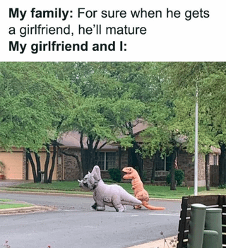 A meme contrasting family expectations of maturity with humorous reality. The top text reads 'My family: For sure when he gets a girlfriend, he'll mature' setting up the expectation of grown-up behavior. The bottom text 'My girlfriend and I:' is followed by an image of two people wearing gray and orange dinosaur costumes walking in a residential neighborhood, subverting the expectation with playful, childlike behavior. The humor arises from the sarcastic contrast between the family's assumption of maturity and the couple's silly, lighthearted actions.