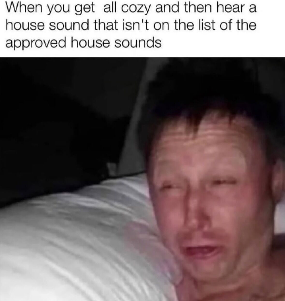 This is a single-panel relatable reaction meme. Text at the top of the image reads "When you get all cozy and then hear a house sound that isn't on the list of the approved house sounds". Below the text is a photograph of a man lying in bed propped against a white pillow, with a disoriented, alarmed, squinting expression that conveys being frozen with mild fear or confusion right after hearing an unexpected, unrecognizable sound in his home. The humor comes from the widely relatable experience of feeling unsettled by unknown noises when you are already settled and comfortable in your living space.