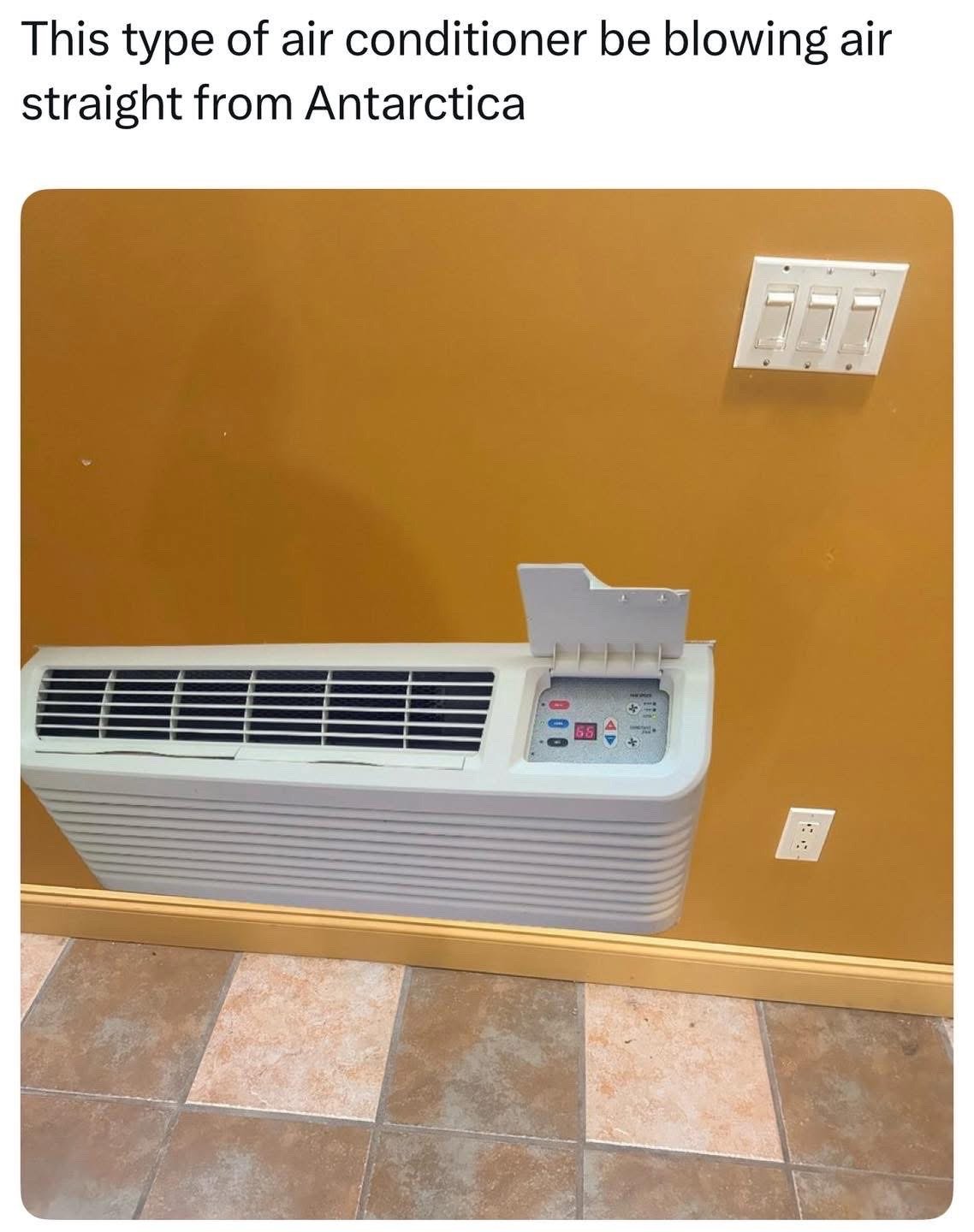 The meme consists of a photograph of a white through-the-wall PTAC (Packaged Terminal Air Conditioner) unit mounted on a mustard yellow interior wall, above square stone tile flooring. A three-switch light panel and standard electrical outlet are visible on the wall next to the AC, whose digital display shows a temperature of 55 degrees Fahrenheit. Black text above the photo reads 'This type of air conditioner be blowing air straight from Antarctica', using humorous hyperbole to joke that these common AC units (often found in hotels, motels, and older multi-unit residences) produce extremely cold air that feels as frigid as the temperatures in Antarctica.
