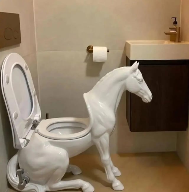 This image displays an absurd, custom-designed functional toilet shaped like a half white horse. The toilet bowl and seat are integrated into the horse's body: the front half of the horse (head, neck, front legs) extends outward from the bowl, while the rear half (hindquarters, hind legs) sits against the bathroom wall connected to the toilet plumbing. It is installed in a modern, clean residential bathroom next to a standard wall-mounted sink and toilet paper holder. The design is highly unusual and surreal, with the original poster asking if anyone wants to own this weird toilet. The humor comes from the completely unnecessary, bizarre artistic design of a common household item.
