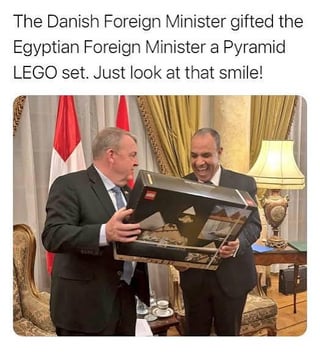 A photograph capturing a friendly diplomatic moment between the Danish and Egyptian Foreign Ministers. The Danish minister, dressed in a dark suit, is holding and presenting a large LEGO box featuring an image of a pyramid. The Egyptian minister, also in a suit, stands beside him, smiling broadly and appearing amused by the gift. The setting is a formal room with elegant curtains, a table lamp, and flags (likely representing Egypt and Denmark) visible in the background. Overlaid text at the top of the image explains the context: 'The Danish Foreign Minister gifted the Egyptian Foreign Minister a Pyramid LEGO set. Just look at that smile!'