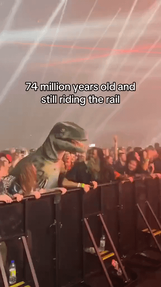 A photograph of a lively concert scene featuring a person dressed in a full T-Rex dinosaur costume leaning on the front barrier ("riding the rail") alongside other concert-goers. The background shows bright stage lights and a dense crowd of fans. Overlaid text at the top of the image reads: "74 million years old and still riding the rail," creating a humorous contrast between the prehistoric dinosaur (which lived approximately 74 million years ago) and the modern concert activity of leaning on the barrier to watch the performance.