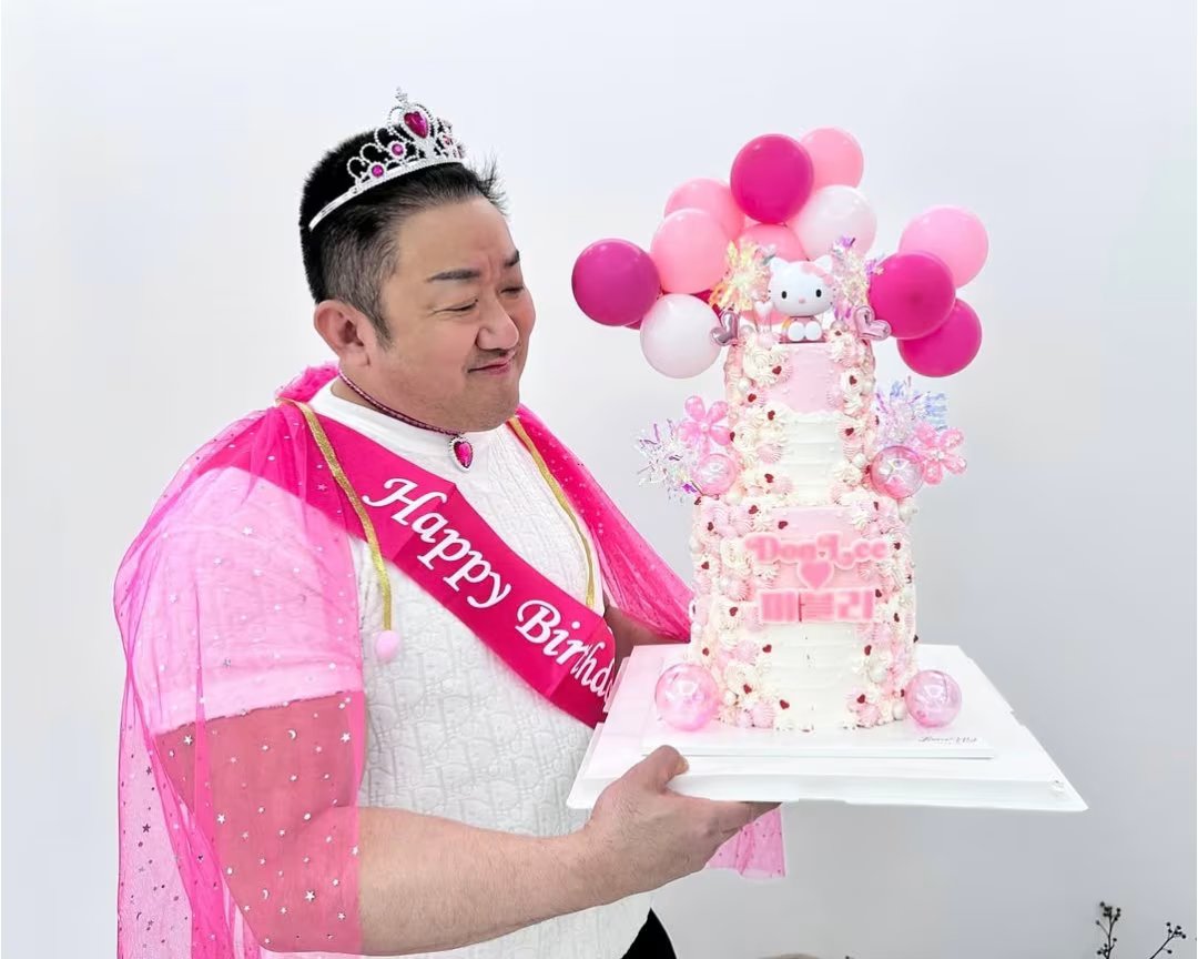 A man dressed in a pink tulle cape, a pink sash inscribed with 'Happy Birthday', and a silver tiara adorned with pink gemstones, holding a tall, elaborately decorated birthday cake. The cake is primarily pink and white, featuring a Hello Kitty figurine on top, surrounded by clusters of pink and white balloons. Additional cake decorations include floral accents, snowflake-like ornaments, and the text 'Dog Love' written in pink icing with a small heart. The man is smiling and appears to be in a cheerful mood, standing against a plain white background that emphasizes the vibrant colors of his outfit and the cake.